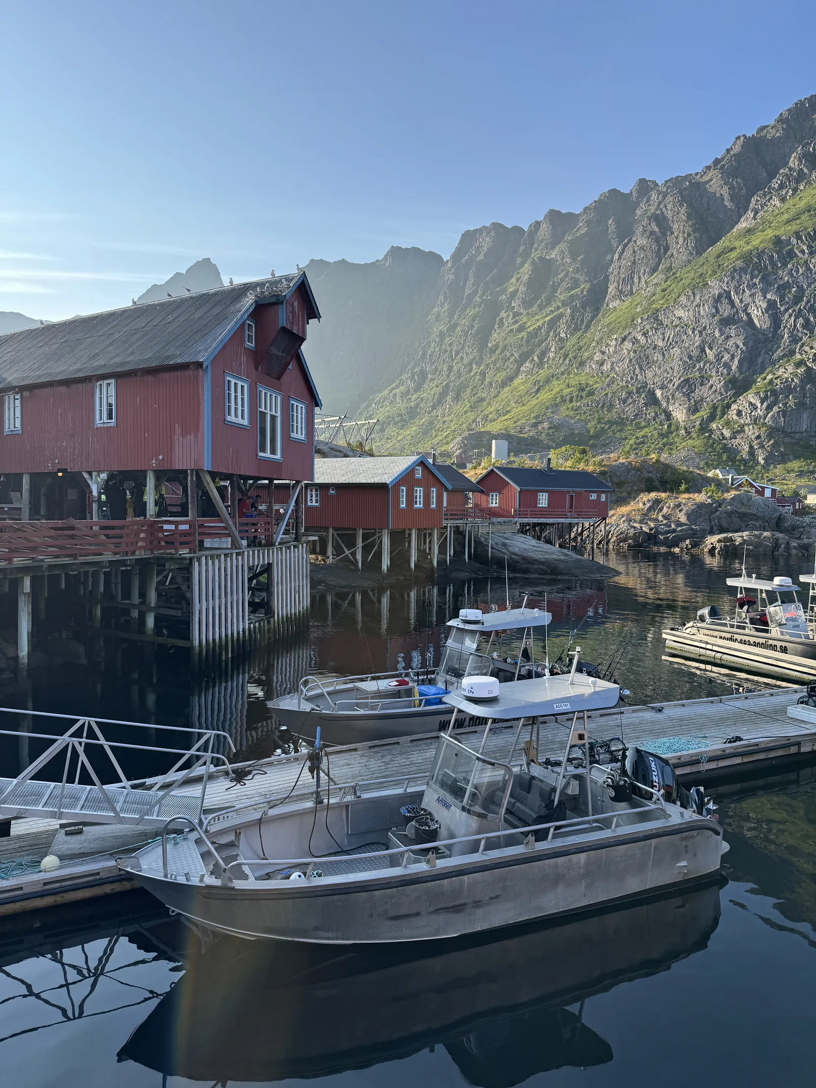 Our photo of the historic red rorbuer of Å standing on stilts over the calm harbour beneath the Lofoten mountains, Norway