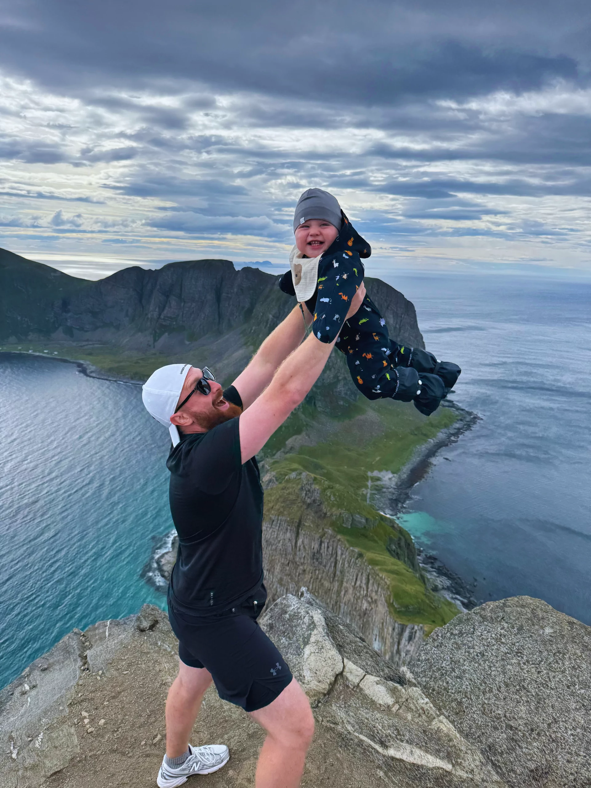 A Gowme.travel author lifting our child at the Håen summit on Værøy, with the Lofoten coast and the Norwegian Sea behind