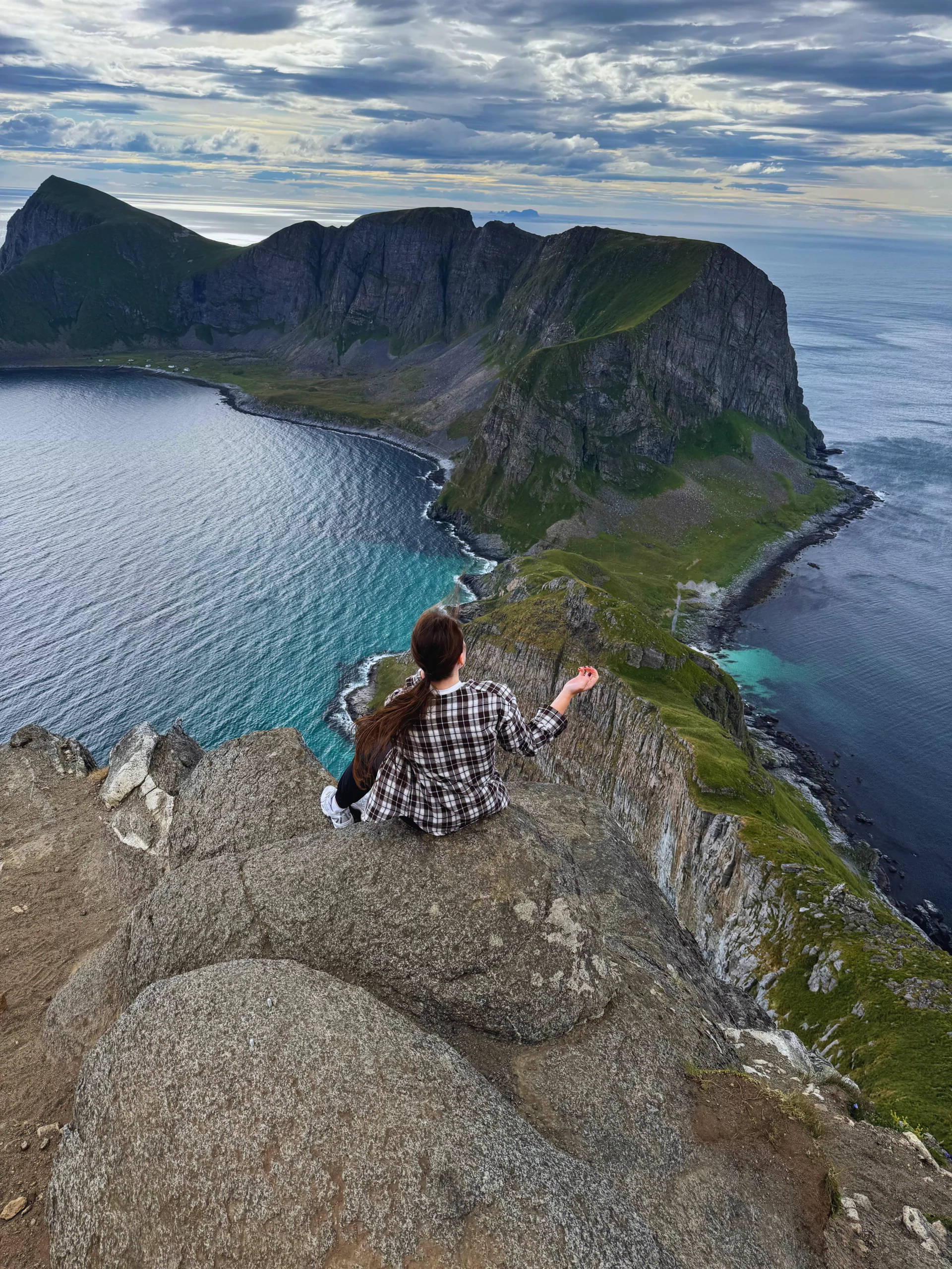 A Gowme.travel author on the cliff edge of the Håen summit, looking out over Værøy's turquoise bays and ridges, Lofoten