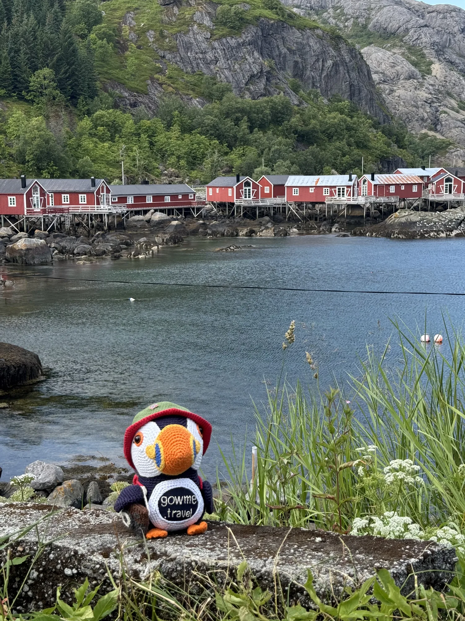 Our Gowme.travel mascot by the water, with Nusfjord's red rorbuer across the harbour, Lofoten